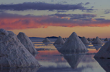 Salar de Uyuni in Bolivien. Durch die Regenzeit ist die Salzwüste mit Wasser bedeckt. Aus dem Wasser ragen kegelförmige Salzformationen. Himmel und Wolken sind durch die Dämmerung in warme Farben getaucht und spiegeln sich im Wasser.